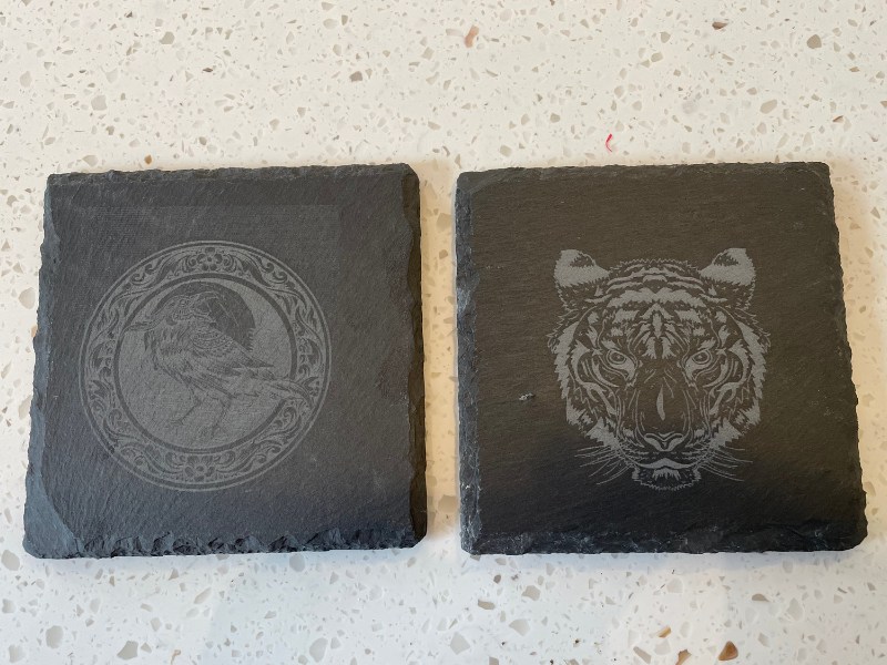 Two laser engraved coasters laying side by side to see the differences in laser settings. One is 12% power and 400mm/s on the left, one is 18% power and 300mm/s power on the right.